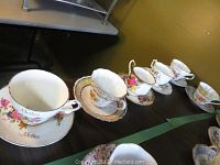 Photo of an assortment of seven teacups and saucers arranged on a table. Includes a 'Mother' labeled set and various floral pattern cups.