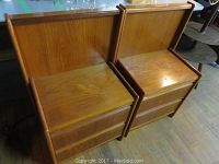 Pair of teak night tables placed side by side. Both show natural wood grain finish with raised back panel and two drawers.