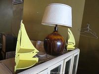 Vintage ceramic lamp with brown glaze and beige fabric shade flanked by two yellow painted wooden corner shelves on top of white cabinet.