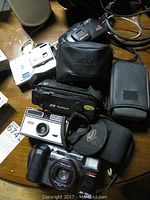 Lot of 7 vintage cameras on wooden table including Kodak Instamatic, Minolta AF-Tele Super, Sony Cyber-shot and Pentax.