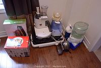 Overview of small kitchen appliances and accessories grouped on floor showing juicer, food processor, cooler box, steamer and humidifier.