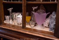 Shelf display containing multiple glassware pieces including a purple frosted glass vase, clear shot glasses, ceramic figure, brass vase, and sterling silver condiment bowl.