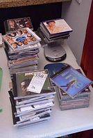 Multiple stacks of assorted music CDs, including various artists and soundtrack albums, arranged on a table alongside two portable CD players, one silver (JVC) and one blue (Optimus Hyper Das).