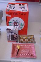 Magic Bullet Deluxe blender box with French press coffee maker set, rectangular decorative plate, and pink ceramic ashtray plate visible on table.