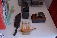 Side view of vintage knife in dark leather sheath, wooden model ship, jewelry box with brass scale, and binoculars case on table