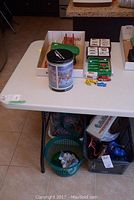 Overview of table with green basket bin underneath, and various boxed or loose items on table including candle warmer and cassette tapes