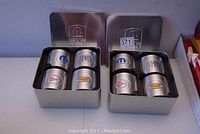 Two open metal tins each holding four metal beer bottle/can insulators, showing automotive logos.