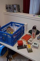 Assorted items on table including blue plastic crate with bubble wrap, measuring spoons, bottles, boxes, badges, and hats