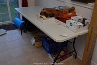 White folding table in a room, top has several items placed including boxes and wooden objects, table measures 72x30x30 inches as given by seller.
