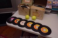 Photo showing several 45 RPM records with orange and pink labels displayed in front of two vintage record storage boxes, along with two tennis balls.