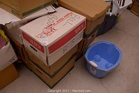 Photo showing stacked cardboard boxes and a blue plastic bucket with a white handle nearby on the floor.