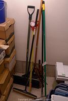 Full view of the garden tools leaning against a wall showing multiple shovels, rakes, brooms and markers.