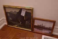 Two framed artworks leaning against wall on wooden floor; larger landscape painting on glass and smaller city skyline print.