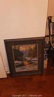 Framed print leaning against wall, depicting a nature scene with a river and trees in sunset/sunrise lighting.
