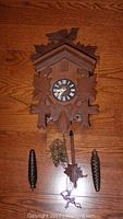 Front view of the wooden cuckoo clock with detached pine cone weights and pendulum laid out on a wood floor.