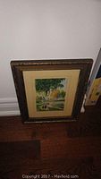 Framed 5x7.5 watercolour painting signed by Ian H Shaw placed against a wall on a wooden floor showing a landscape with a birch tree, water and houses.