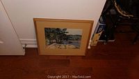 Watercolour painting leaning against a wall on hardwood floor, showing the full framed artwork with a visible natural landscape scene.