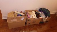 Three boxes filled with various sewing supplies and fabric on the floor against a white wall.