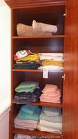 Shelved linen items folded and stacked including beige rolled towels or bath mats top shelf, assorted colored towels and tablecloths on second shelf, folded towels and linens in green, pink, and blue on lower shelves.