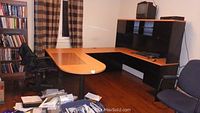 Front view of L-shaped Belair computer desk set in a room, showing wood and black finishes, upper hutch, and workspace area with chairs and bookshelves.