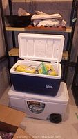 Blue Igloo cooler open showing freezer packs and disposable cutlery inside, on top of white Coleman wheeled cooler.