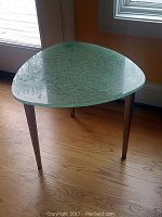 Photo showing the triangular textured glass top and metal legs of the side table, positioned by a window on a hardwood floor.