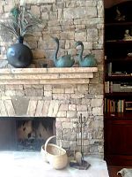 Wide view of stone fireplace mantle with two metal duck planters, black round vase with silk feathers, woven basket on hearth, and metal fireplace tools.