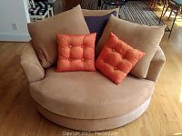 Front view of large circular beige upholstered chair with two orange and two beige cushions