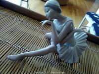 Side view of glossy porcelain ballerina figurine seated with one leg extended and one arm raised near face, showing detailed facial features and tutu skirt.