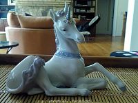 Front and side view of a white porcelain unicorn figurine with lavender mane and tail, blue collar, and a floral horn. The unicorn is seated with front legs raised.