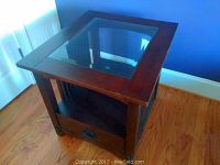 Wood side table with square glass inset top shown from an angle, placed on hardwood floor near window and purple wall.