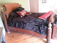 Photo showing full wooden bed frame with detailed carved posts, upholstered headboard and footboard, dressed with black quilted bedding and decorative pillows.