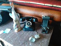 Wide view showing black rotary phone, Kodex Kodak camera, two pocket watches, and ceramic bird figurine on a marble-like surface.