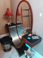 Vanity desk with large oval wooden framed mirror, glass top, and curved metallic base, alongside a round cushioned stool with metallic base. Items are placed beside a unique red lamp.