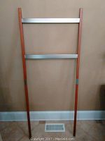 Photo of a tall towel rack with wooden sides and horizontal metal bars against a beige wall.