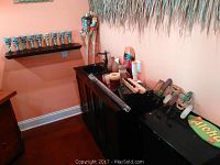 Tiki bar counter with skull shot glasses on display rack, tiki torches, wooden paddle decorations, tiki bar roof thatching, and green 'Aloha' sign on black fridge or cooler.