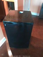 Full front view of the GE black mini refrigerator, showing smooth top and door with GE logo.