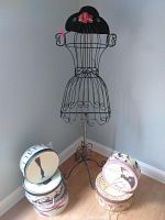 Front view of black wire dress form mannequin with decorative black hat featuring pink flowers and five assorted decorative hat boxes stacked beside it.