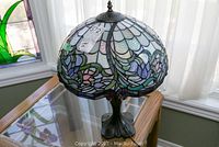 Tiffany style table lamp showing floral leaded glass shade and bronze metal base on glass table near window.