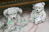 Two concrete bear figures displayed on an indoor floor surface, one textured shaggy bear and one dressed bear with hat and jacket, both tagged with green labels.