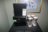 Photo showing 3 black boxes stacked next to 4 goblets on black surface, with artwork in background.