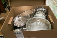 View inside cardboard box showing various silverplate trays and servers, including a scalloped edge tray and a covered glass dish with patterned bottom.