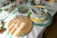 Table display showing stack of clear Cornflower glass side plates, colorful Joie Vivre MSC plates, cocktail plates in container, ceramic serving tray, and wooden cheese board with glass dome.