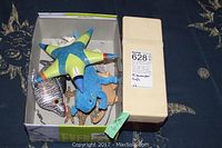 View of the box containing assorted plush toys including a star-shaped fabric toy with clown face, a silver fish, a blue bear, and other small stuffed animals.