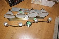 Photo of two wall mount light strips on a wooden floor, one with three frosted glass bell shades on a brass-toned bar, the other a curved metal bar with four small spotlight bulbs.