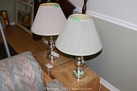 Pair of silver-tone table lamps with pleated white shades on small wooden tables next to a patterned sofa, lamps are turned off, showing condition and styling.