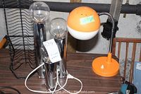 Two cylinder chrome or metallic desk lamps with round bulbs, and one orange and white desk lamp with flexible neck on table.
