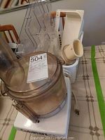 Photo showing food processor main bowl, lid, and attached components on top of white base with instruction sticker visible.