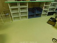 Photo showing multiple white and blue plastic wire mesh storage containers stacked on the floor along a wall.