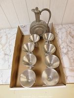 Pewter decanter and eight metal glasses arranged in a box, showing the decanter's round body and stopper with glasses lined up in two rows.